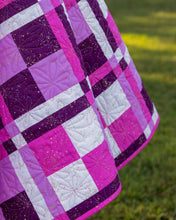Load image into Gallery viewer, A close-up of Windows Party Quilt featuring a pink, purple, and white plaid pattern with floral quilting and subtle gold flecks, set against a blurred green grass background.