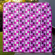 Load image into Gallery viewer, A square quilt with a pink, purple, and white checkerboard block pattern draped outdoors on a concrete path next to a gray metal building and green grass.