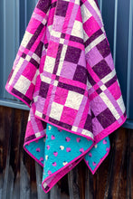 Load image into Gallery viewer, A handmade quilted blanket with a pink, purple, and white plaid pattern on the front, and a turquoise reverse side with a small heart print, hanging against a dark wooden wall.