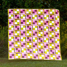 Load image into Gallery viewer, A square quilted blanket with a colorful geometric pattern of pink, purple, yellow, and white squares, displayed outdoors on a grassy lawn with green bushes in the background.