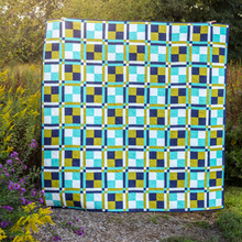Load image into Gallery viewer, A square quilted blanket or quilt with a geometric patchwork pattern in navy blue, teal, lime green, and white, displayed outdoors on a gravel path surrounded by green foliage and yellow flowers.