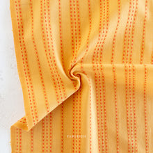 Load image into Gallery viewer, Close-up of orange fabric with subtle pattern. Embrace the charm of the rainbow with Fableism Supply Co’s ‘Lucky Loom’ fabric collection—a dazzling fusion of mixed weight wovens inspired by moments of serendipity. This collection’s palette radiates warmth and whimsy, featuring golden yellows, soft pinks, earthy greens, tranquil blues, and rich purples, each reminiscent of the colors in a lucky rainbow. Available at globalfibershop.com.