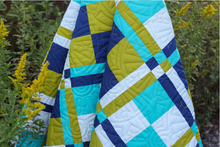 Load image into Gallery viewer, A close-up of a quilted patchwork blanket draped outdoors among green plants and yellow wildflowers. The blanket features a geometric pattern of navy blue, turquoise, olive green, and white stitched sections.