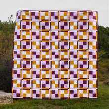 Load image into Gallery viewer, A large quilted patchwork throw with a geometric checkered pattern in purple, white, and mustard yellow, displayed outdoors on a grassy lawn against a cloudy sky.