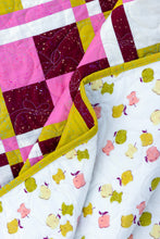 Load image into Gallery viewer, Close-up of a quilted fabric with a multicolor patchwork design in pink, burgundy, yellow, and white, folded over to show a white backside printed with colorful cartoon fruit characters.

