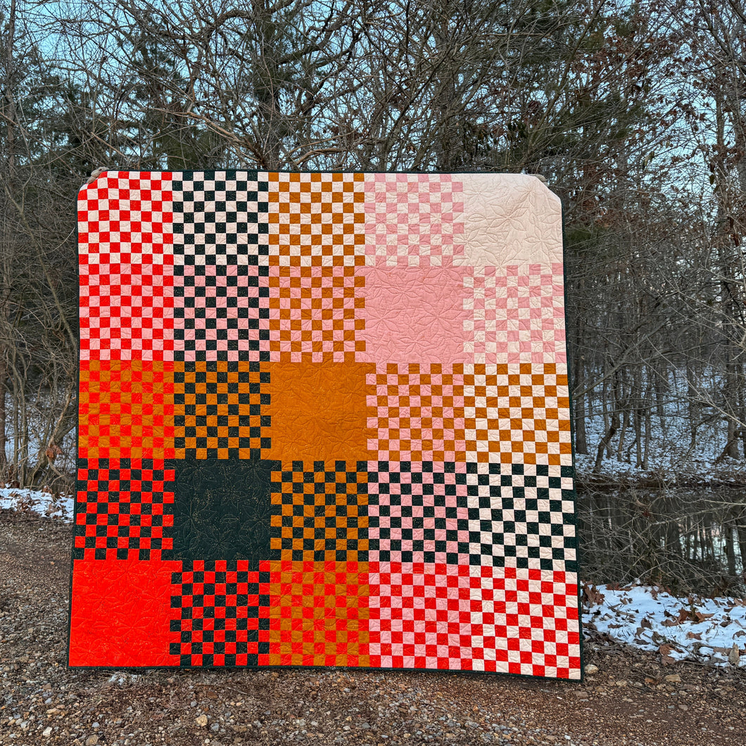 Checkered quilt with orange, black, and pink pattern on a gravel path with trees in the background. Norma Jean bundle available at globalfibershop.com.