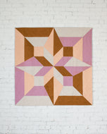 Geometric quilt with pink, brown, and beige colors on a white brick wall background. Arete Quilt bundle by Eudaimonia Studio available at globalfibershop.com.