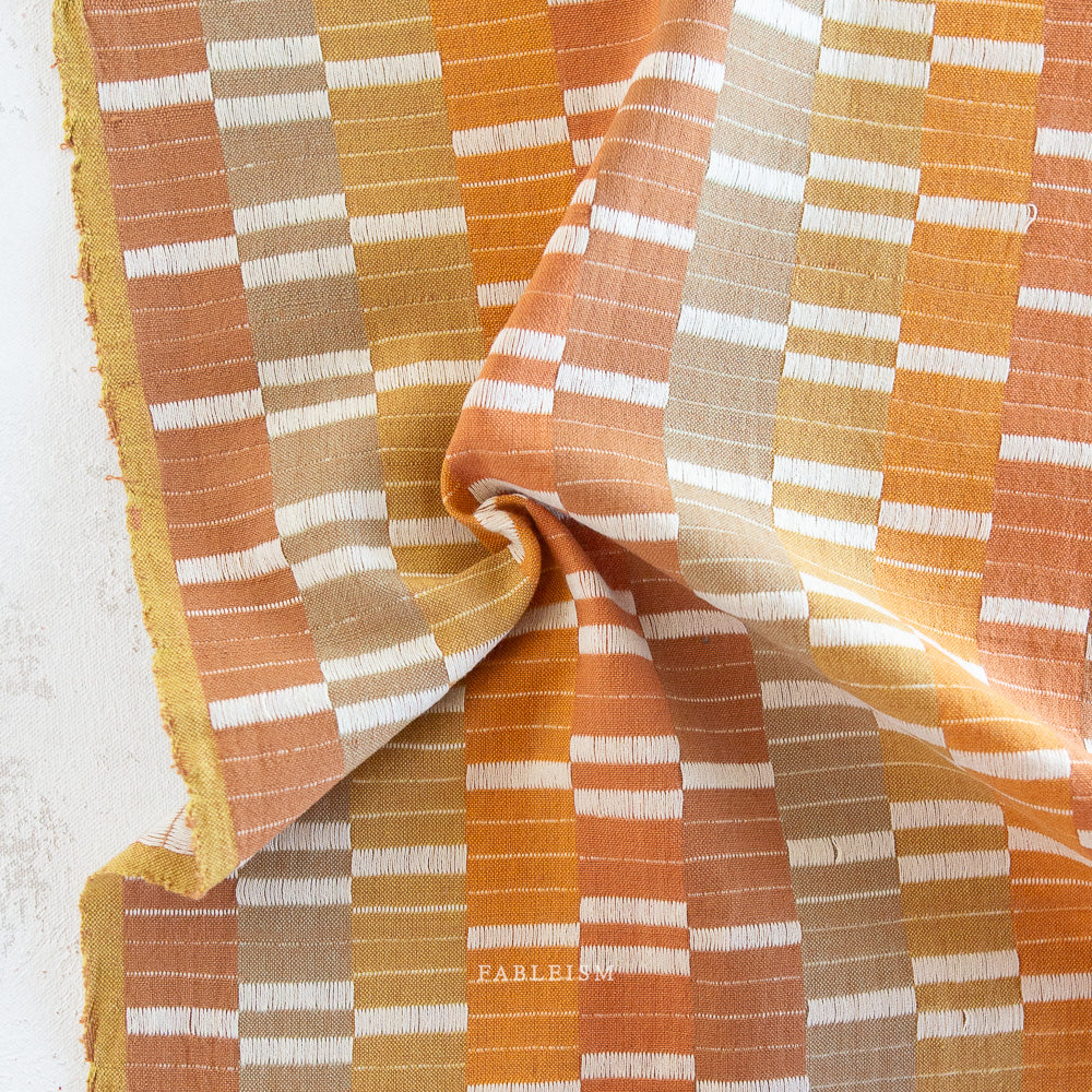 Textured fabric with orange, brown, and white stripes. Embrace the charm of the rainbow with Fableism Supply Co’s ‘Lucky Loom’ fabric collection—a dazzling fusion of mixed weight wovens inspired by moments of serendipity. This collection’s palette radiates warmth and whimsy, featuring golden yellows, soft pinks, earthy greens, tranquil blues, and rich purples, each reminiscent of the colors in a lucky rainbow. Available at globalfibershop.com.