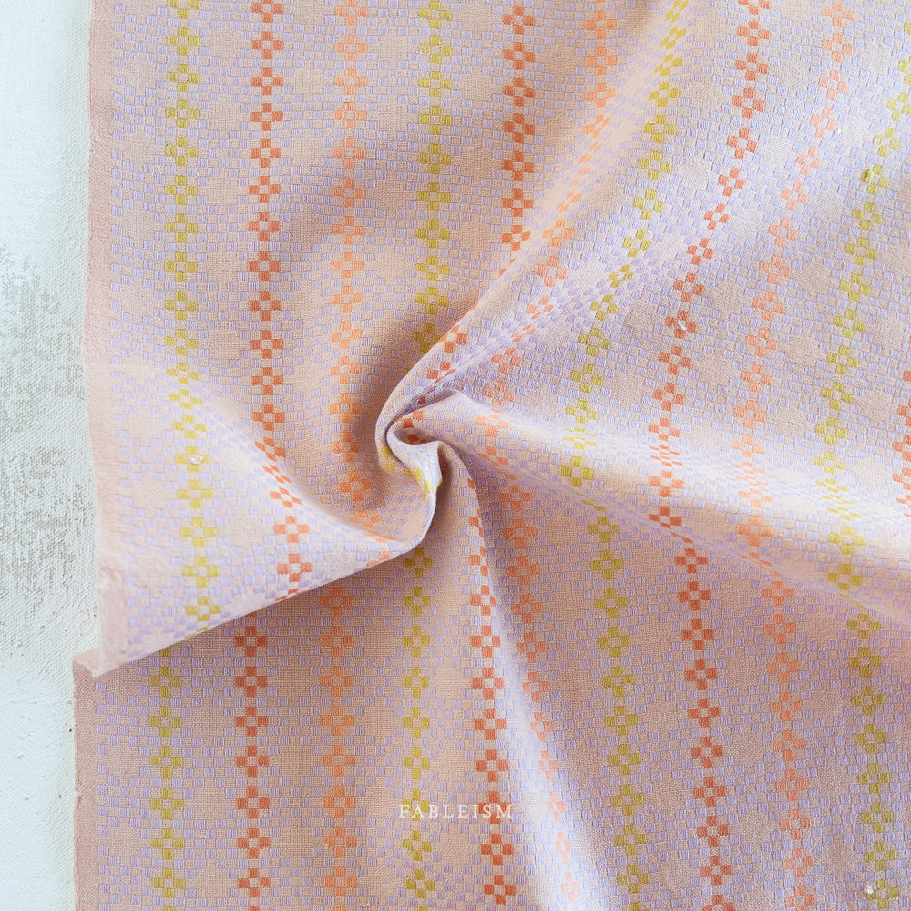 Fabric with a geometric pattern on a light background.  Embrace the charm of the rainbow with Fableism Supply Co’s ‘Lucky Loom’ fabric collection—a dazzling fusion of mixed weight wovens inspired by moments of serendipity. This collection’s palette radiates warmth and whimsy, featuring golden yellows, soft pinks, earthy greens, tranquil blues, and rich purples, each reminiscent of the colors in a lucky rainbow. Available at globalfibershop.com.