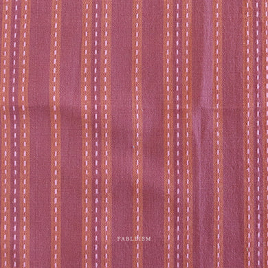 Textured fabric with vertical stripes in shades of purple and brown on a matching background.  Embrace the charm of the rainbow with Fableism Supply Co’s ‘Lucky Loom’ fabric collection—a dazzling fusion of mixed weight wovens inspired by moments of serendipity. This collection’s palette radiates warmth and whimsy, featuring golden yellows, soft pinks, earthy greens, tranquil blues, and rich purples, each reminiscent of the colors in a lucky rainbow. Available at globalfibershop.com.