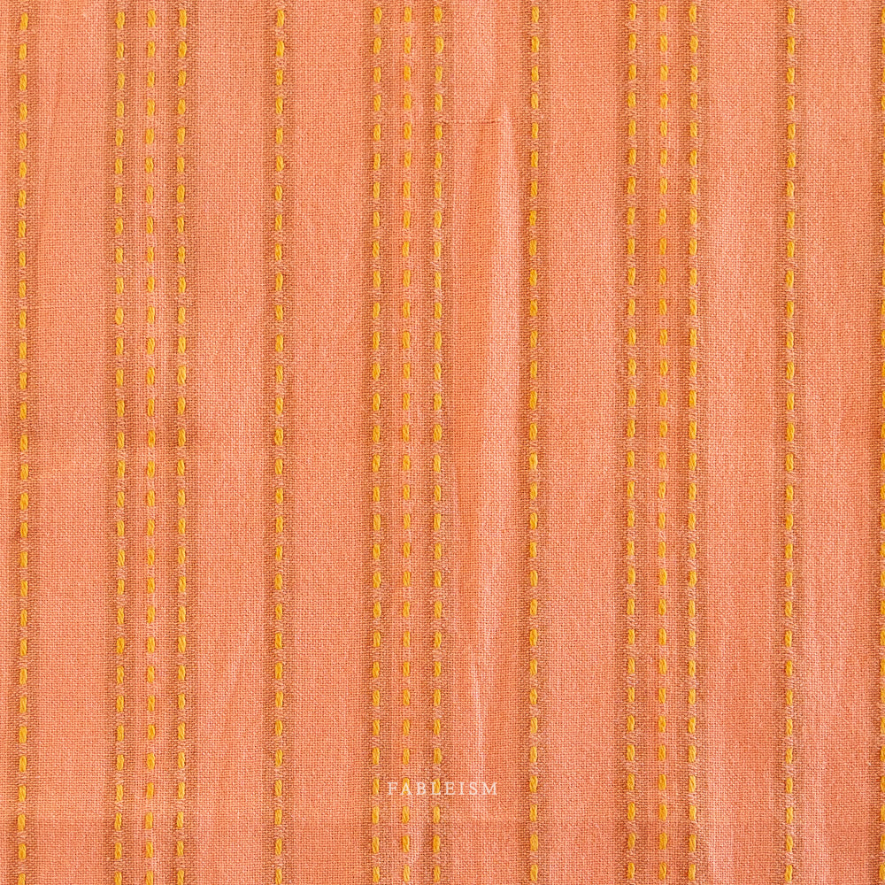 Woven fabric with vertical yellow stitching on a peach background.  Embrace the charm of the rainbow with Fableism Supply Co’s ‘Lucky Loom’ fabric collection—a dazzling fusion of mixed weight wovens inspired by moments of serendipity. This collection’s palette radiates warmth and whimsy, featuring golden yellows, soft pinks, earthy greens, tranquil blues, and rich purples, each reminiscent of the colors in a lucky rainbow. Available at globalfibershop.com.