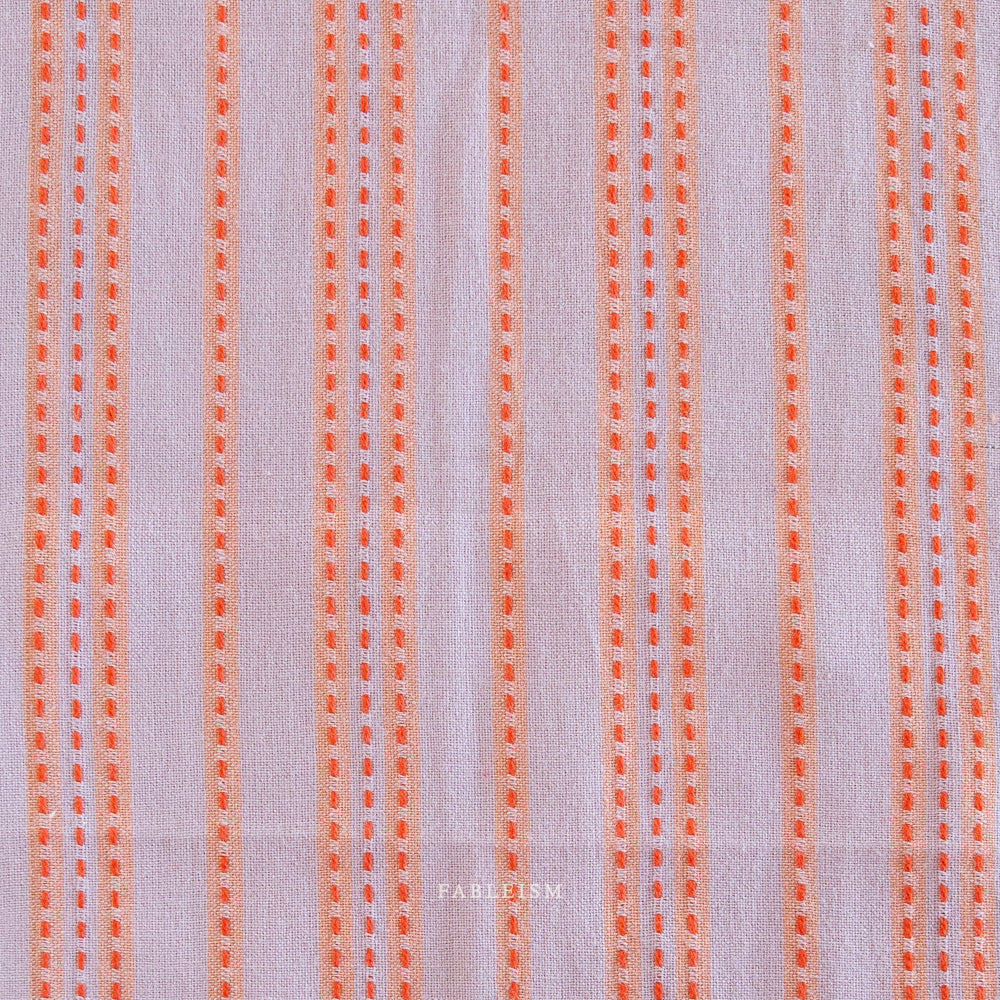 Woven Fabric with orange vertical stripes on a light lavender background.  Embrace the charm of the rainbow with Fableism Supply Co’s ‘Lucky Loom’ fabric collection—a dazzling fusion of mixed weight wovens inspired by moments of serendipity. This collection’s palette radiates warmth and whimsy, featuring golden yellows, soft pinks, earthy greens, tranquil blues, and rich purples, each reminiscent of the colors in a lucky rainbow. Available at globalfibershop.com.