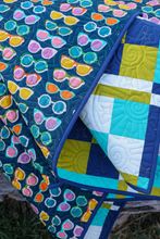 Load image into Gallery viewer, A close-up of a colorful quilted blanket or throw featuring a repeating pattern of colorful sunglasses on a dark blue background, with a folded section revealing a patchwork design in blue, yellow, white, and turquoise.
