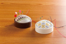 Load image into Gallery viewer, Two round pincushions on a wooden tabletop. The left pincushion is dark brown with a white cross-stitch pattern and multicolored pins.
